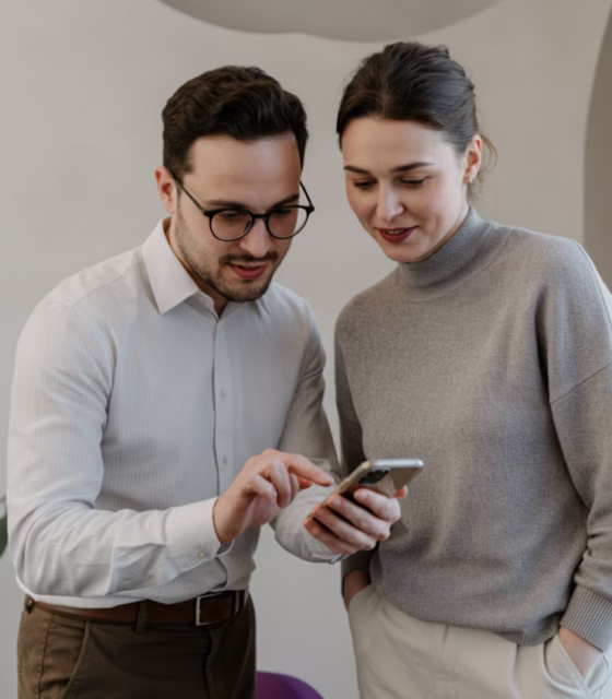 A man wearing glasses and a white shirt shows something on his phone to a woman in a gray sweater who is looking at the screen.