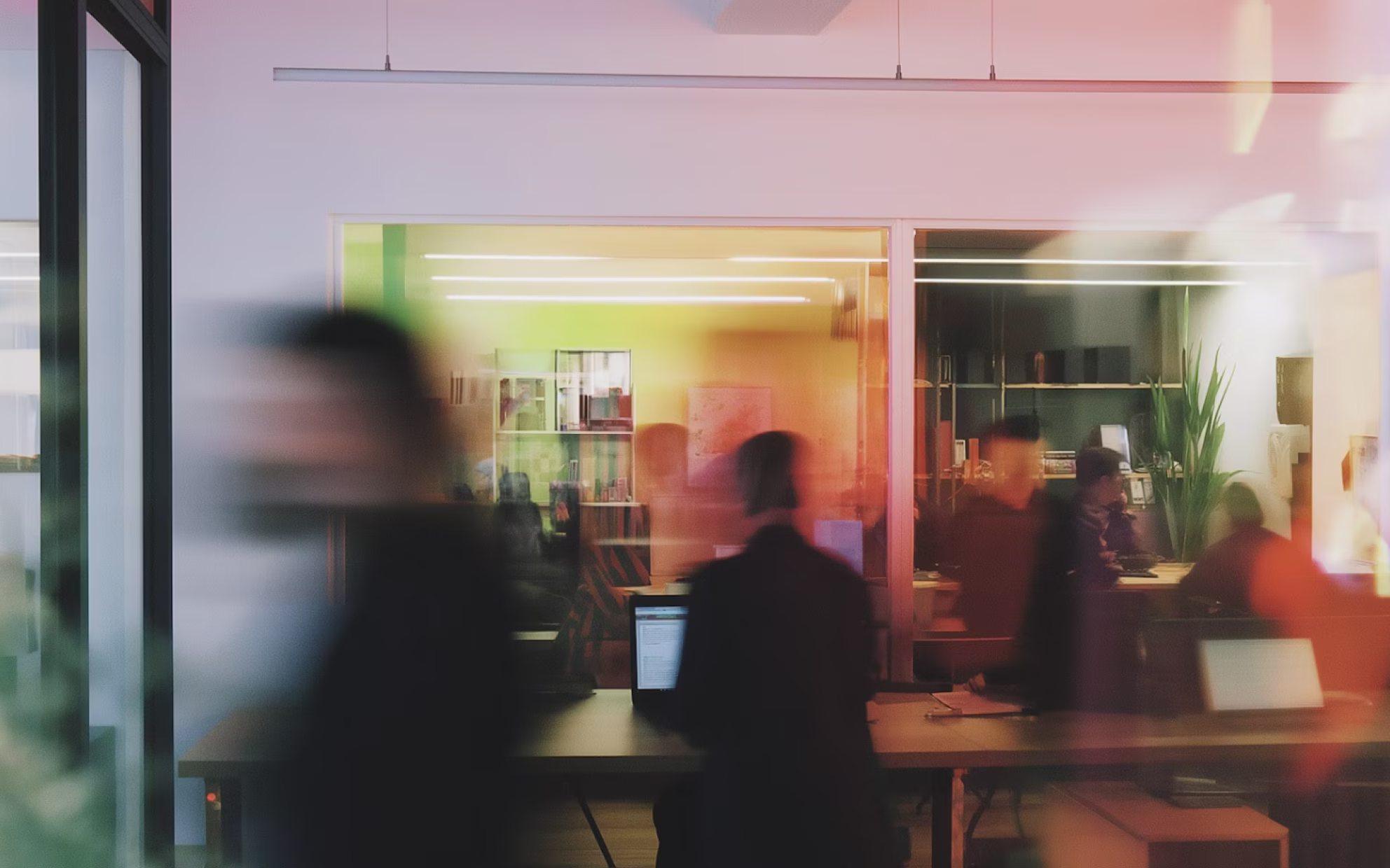 Blurred silhouettes of people working in a modern office with laptops and glass partitions.