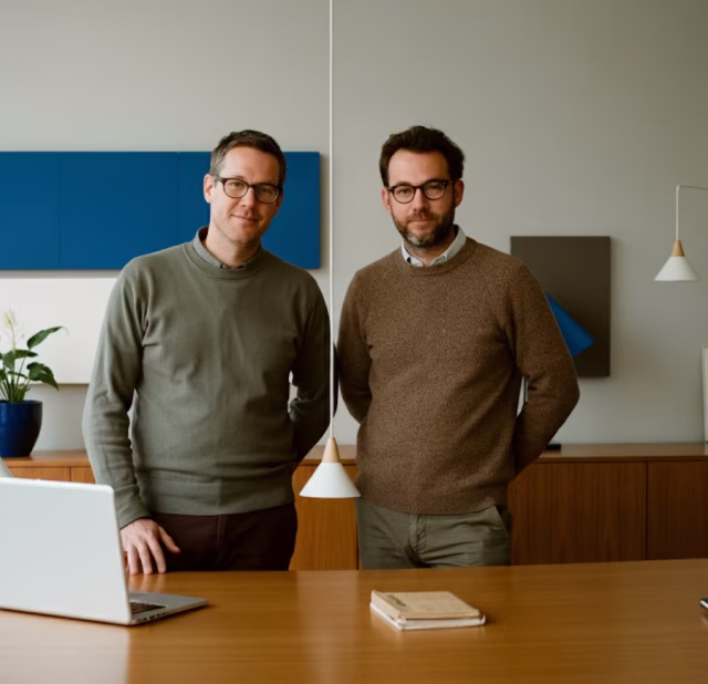 Two men wearing glasses and sweaters stand behind a wooden table with a laptop and notebook in a modern office.