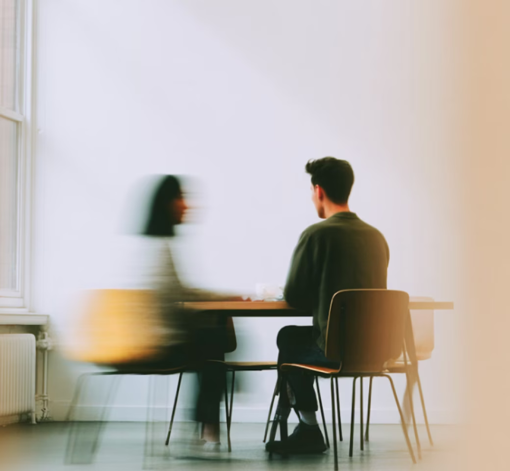 Two people sitting across from each other at a table in a minimalist room with a blurred effect on one person.